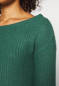 Grüner gestrickter Pullover mit weitem, geripptem Ausschnitt und strukturiertem Muster. Der Stoff wirkt weich und der Schnitt locker.