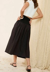 Mustapohja, virtaava maksihame, jossa on kerrostettu vyötärö. Tyylitelty istuvalla valkoisella paidalla ja mustilla sandaaleilla teksturoidulla lattialla.