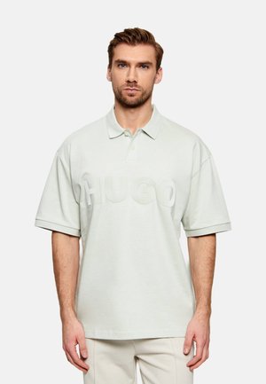 Hombre con pelo castaño claro y barba que lleva una camisa polo blanca extragrande con grandes letras "HUGO" en relieve en la parte delantera.