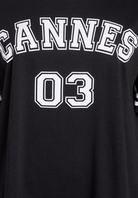 Melns T-krekls ar uzrakstu "CANNES" un "03", kas drukāts treknajos baltajos blokos. Īsām piedurknēm ar svītrām; izgatavots no mīksta kokvilnas materiāla.