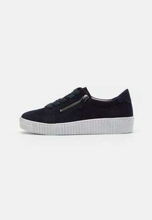 Baskets en suède noir avec une semelle en caoutchouc texturé blanche. Comprend des lacets et un détail de fermeture éclair sur le côté.