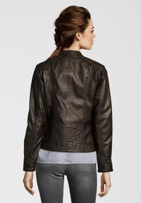 Dunkelbraune Lederjacke mit taillierter Form, sichtbaren Steppnähten und Stehkragen. Über einem hellgrauen Oberteil und schmal geschnittenen Jeans getragen.