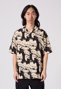 Short-sleeve button-up shirt in beige and black, featuring a striking abstract face pattern, lightweight fabric, and a classic collar.