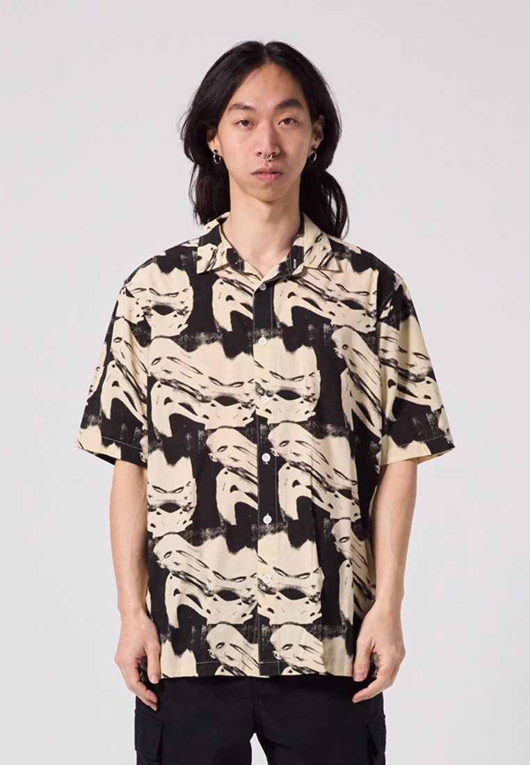 Short-sleeve button-up shirt in beige and black, featuring a striking abstract face pattern, lightweight fabric, and a classic collar.
