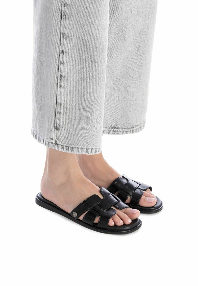 Sandalias deslizantes de cuero negro con diseño de tiras trenzadas, suela plana y punta abierta, combinadas con jeans de pierna ancha de color gris claro.
