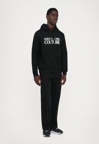 Νεαρός άνδρας στέκεται φορώντας μαύρη κουκούλα με κείμενο "Versace Jeans Couture", μαύρο παντελόνι και μαύρα sneakers σε απλή λευκή γαλάζια.