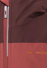 Rote und kastanienfarbene Jacke mit einem Reißverschluss vorne. Der Stoff ist glatt und hat ein gesticktes Logo. Zwei Farben bilden ein Farbblockdesign.