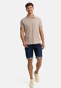 Beige Baumwoll-Polohemd, dunkle Jeansshorts mit umgeschlagenen Säumen und hellfarbige Sneakers, getragen von einem lächelnden männlichen Model.