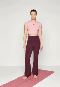 Jauna sieviete stāv basām kājām uz rozā jogas paklāja, ģērbusies rozā topā ar īsām piedurknēm un bordo platās biksēs pret vienkāršu fona.