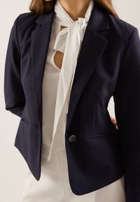 Blazer azul marino de corte entallado, con solapas con muesca, cierre de un solo botón y dos bolsillos laterales. Llevado sobre una blusa blanca con lazo.