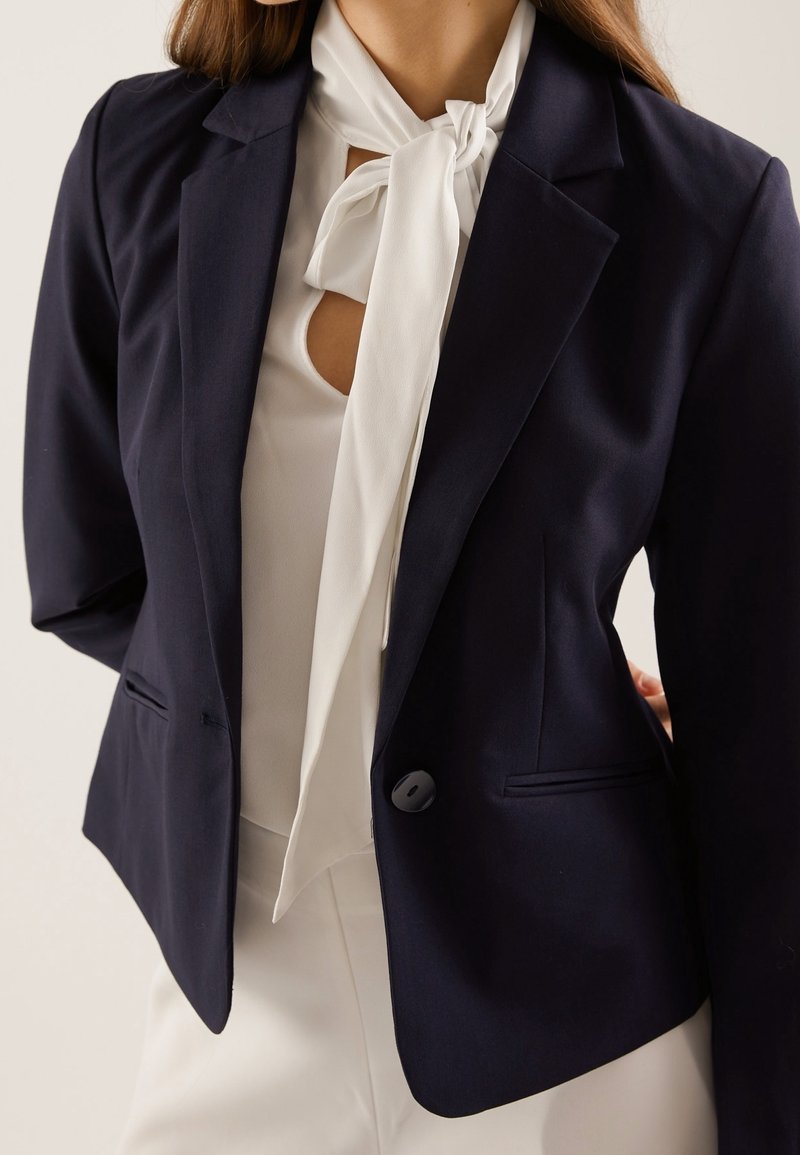 Blazer azul marino de corte entallado, con solapas con muesca, cierre de un solo botón y dos bolsillos laterales. Llevado sobre una blusa blanca con lazo.