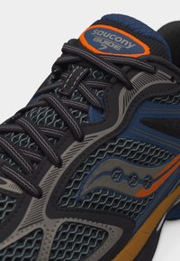 Κοντινό πλάνο παπουτσιού τρεξίματος Saucony Guide 7 με μαύρα κορδόνια, μπλε και πορτοκαλί λεπτομέρειες, και συνδυασμό από δίχτυ και συνθετικά επικάλυψεων.