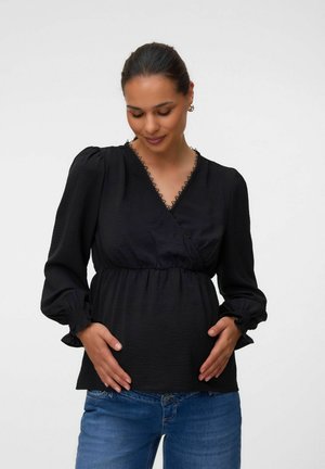 Gravid kvinde iført sort langærmet bluse og blå jeans, der forsigtigt holder sin mave, mens hun blidt kigger nedad mod en ensfarvet baggrund.