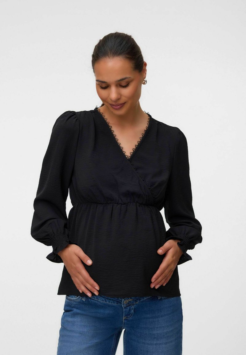 Femme enceinte en blouse noire à manches longues et jean bleu tenant doucement son ventre, regardant doucement vers le bas sur un fond uni.