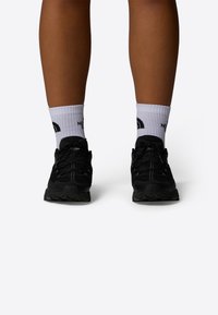 Zapatillas deportivas negras con una superficie texturizada, cierre de cordones y suelas de goma, llevadas con calcetines blancos hasta el tobillo que muestran un logo.