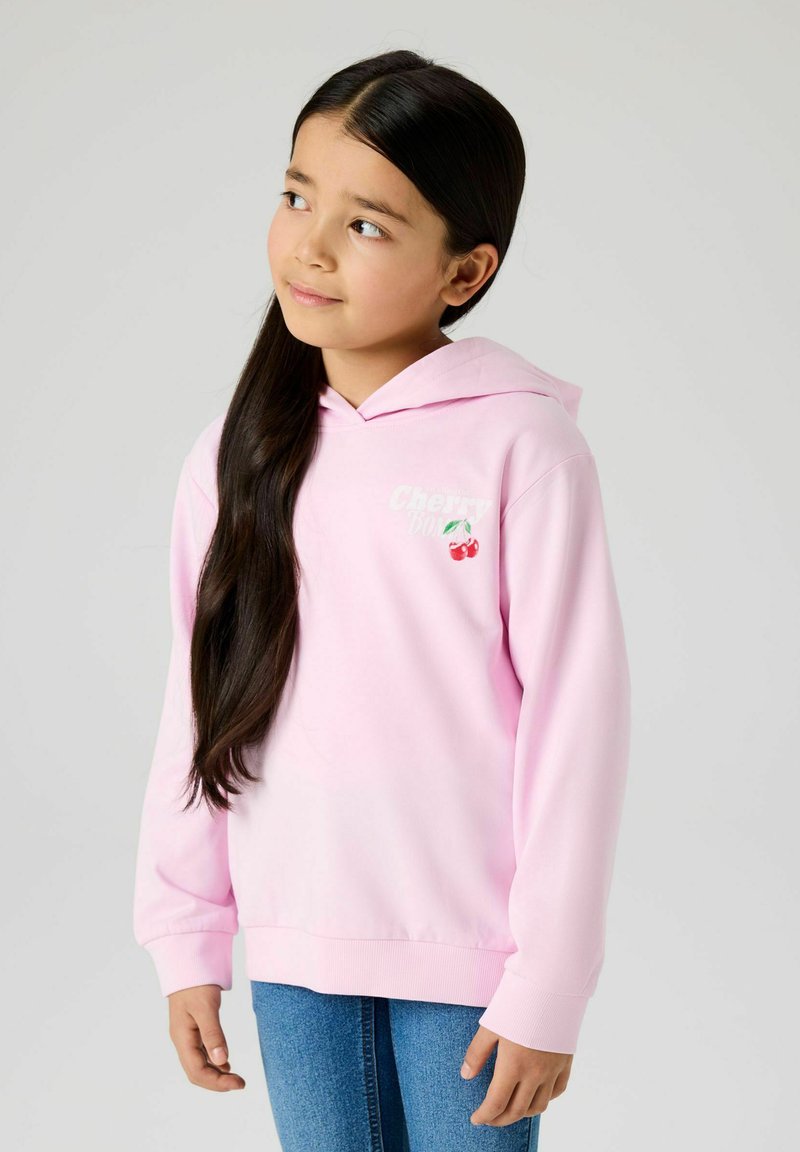 Jeune fille aux longs cheveux foncés, portant un sweat à capuche rose clair avec un motif cerise et un jean bleu, regardant légèrement vers le haut et à droite.