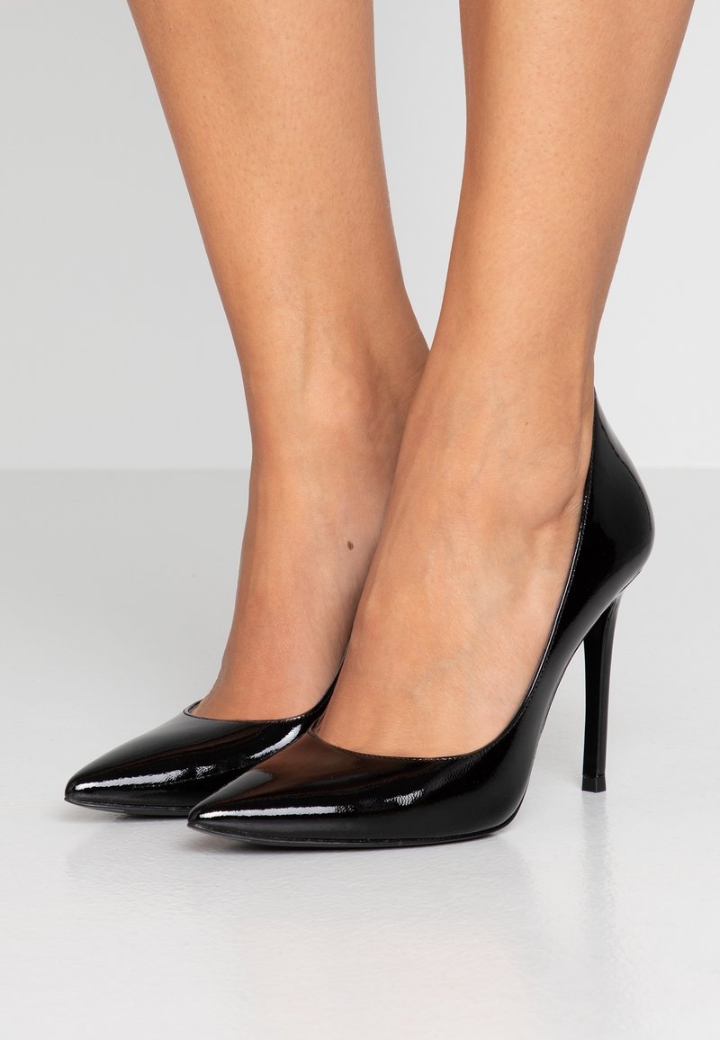 Jambes portant des escarpins à talons aiguilles noirs brillants et pointus sur une surface blanche lisse avec un fond gris uni.