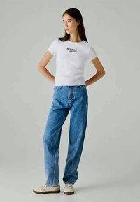 T-shirt branca de manga curta com o estampado "RED BULL RACING", combinada com jeans de ganga azul de corte largo e sapatos beije. Tecido suave, estilo casual.