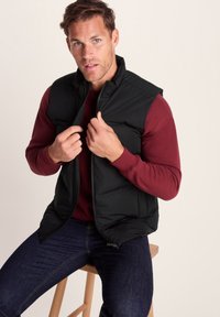Black puffer vest with a zippered front, worn over a burgundy long-sleeve shirt. Model is seated, holding the vest closed with both hands.