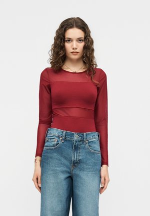 Ung kvinde med krøllet hår iført en tætsiddende rød langærmet top og løse blå jeans, står mod en ensfarvet hvid baggrund.