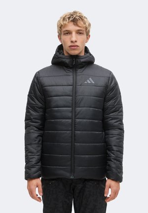 Junge Person mit kurzen lockigen Haaren trägt eine schwarze Steppjacke mit Kapuze und Reißverschluss vorne und steht vor einem einfarbigen hellen Hintergrund.
