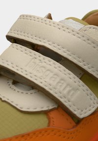 Κοντινή λήψη μπεζ δερμάτινων λουριών βέλκρο σε κίτρινο και πορτοκαλί αθλητικό παπούτσι με ορατή ραφή και ανάγλυφο όνομα μάρκας.