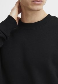 Sudadera negra con cuello redondo, puños acanalados y una textura suave, mostrando un primer plano del escote y parte de la manga.
