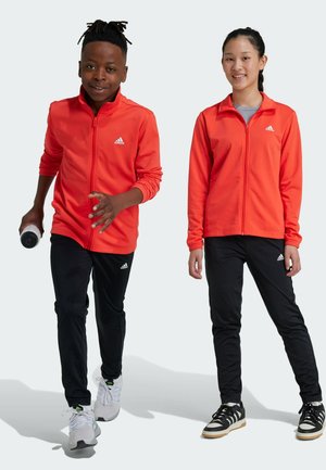 Giubbotti rossi con zip e pantaloni sportivi neri, con accenti del logo Adidas bianco. Il modello a sinistra tiene una bottiglia d'acqua nera.