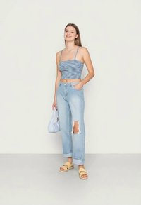 Top cropped azul com riscas, jeans claros desgastados com joelhos desfiados, sandálias amarelas e uma mala branca. Conjunto simples e casual.