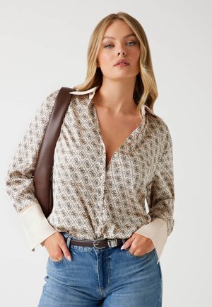 Cream blouse with a geometric brown pattern, featuring a button-up front and white cuffs. Worn with blue jeans and a brown belt.