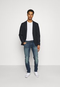 Blazer escuro sobre uma t-shirt branca, combinado com jeans desgastados de um azul claro e ténis brancos. Design simples com corte clássico.