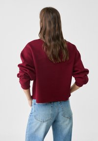 Persona con lunghi capelli castani che indossa un ampio sudore bordeaux e jeans chiari a vita alta, in piedi con le mani in tasca, di spalle.