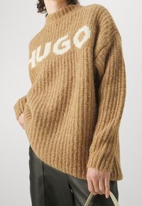 Pull en maille côtelée marron avec un col haut et une coupe oversize, présentant le mot "HUGO" en blanc sur la poitrine. Équilibré avec des manches amples.