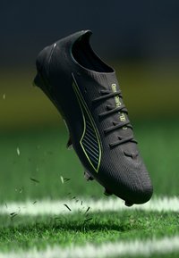 Une chaussure de football noire avec une surface texturée, des accents verts et six œillets pour les lacets, suspendue au-dessus d'un terrain herbeux avec des débris de gazon.