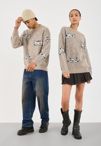 Beiger Strickpullover mit Katzenmotiven, getragen von zwei Models. Eine kombiniert ihn mit blauen Jeans, die andere mit einem schwarzen Rock und kniehohen Stiefeln.