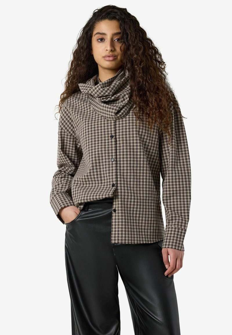 Femme aux longs cheveux bouclés portant une chemise marron à carreaux boutonnée avec un col montant et un pantalon en cuir noir, debout, une main dans la poche.