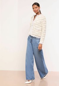 Balta teksturēta jakas ar pūkainu apdari, atvērta priekšpuse, saskaņota ar gaiši zilām platajām biksēm un vērstām baltām papēžiem. Gludas virsmas, ikdienas stils.