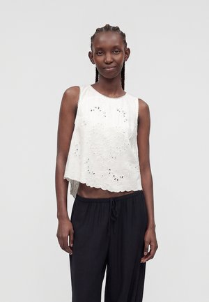 Jeune femme aux cheveux tressés portant un haut blanc sans manches en œillet et un pantalon noir ample, debout devant un fond clair uni.