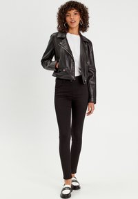 Veste de moto noire en cuir synthétique au design court, avec des zips argentés et des boutons pression, assortie à un jean slim noir et des chaussures noires.