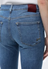 Gros plan d'une personne portant un jean denim bleu avec un patch en cuir marron de la marque et un logo brodé sur la poche arrière.