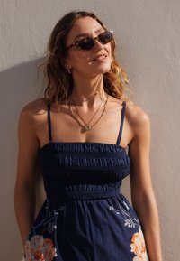 Rochie vaporoasă albastru marin, plisată, cu imprimeu floral și bretele subțiri; modelul poartă ochelari de soare cu ramă de tartar și coliere aurii suprapuse.