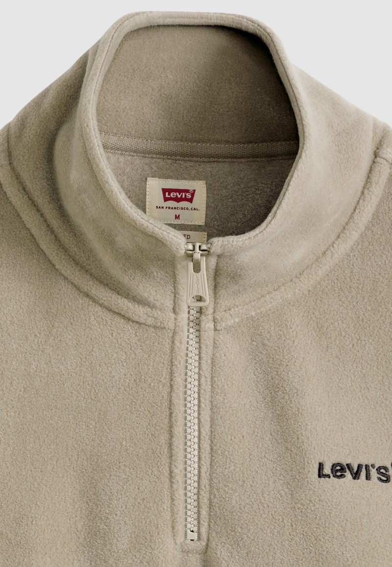 Ljusbeige fleece-tröja med kvartsglipa, hög krage, silverfärgad dragkedja och broderad svart "Levi's"-logotyp på vänster bröst.