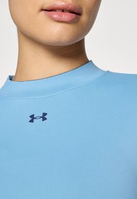 Lys blå, ribbet stoff med høy hals, med en mørk blå Under Armour-logo på brystet. Glatt tekstur og tettsittende design.