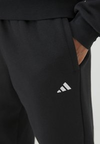 Μαύρες φόρμες από μαλακό ύφασμα, με πλαϊνή τσέπη και λευκό λογότυπο Adidas στο κάτω μέρος του ποδιού. Απλός σχεδιασμός με ελαστική μέση.