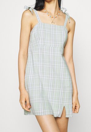 Mujer con vestido sin mangas de cuadros verde claro con tirantes de lazo y una abertura frontal, complementado con collares de cuentas de colores en capas.