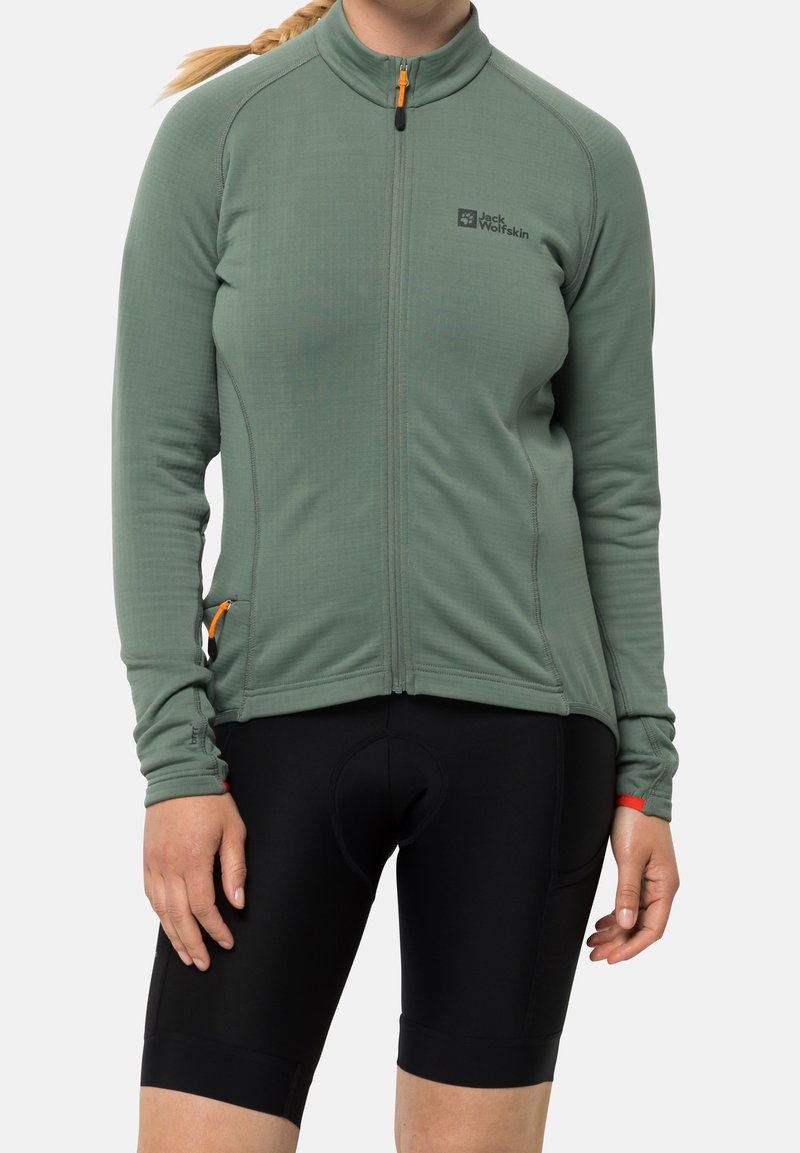 Giacca verde a maniche lunghe con zip realizzata in tessuto testurizzato, tasca laterale con zip arancione, abbinata a pantaloni ciclistici neri.