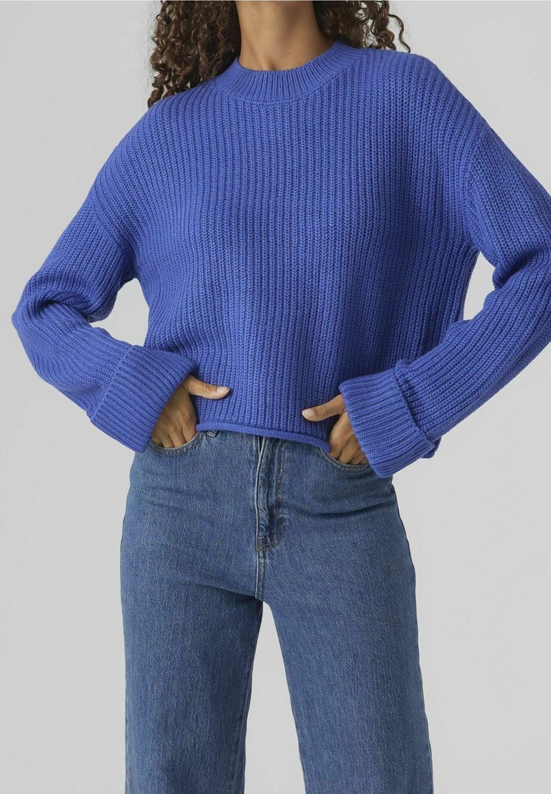 Pull en tricot bleu avec une texture côtelée, un col rond et des poignets larges, associé à un jean taille haute de couleur denim bleu clair.