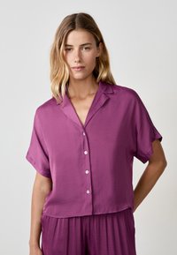 Camicia a maniche corte con bottoni in una vivace miscela di seta viola, caratterizzata da una vestibilità rilassata, colletto a revers e un orlo cropped con una morbida texture.
