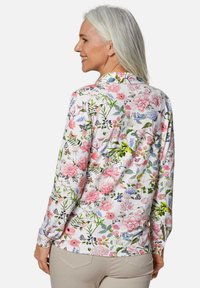 Bluse mit langen Ärmeln im Blumenmuster, die eine Vielzahl farbiger Blumen und Schmetterlinge auf einem weißen Hintergrund zeigt; weicher Stoff mit klassischem Kragen.
