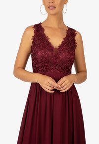 Burgunderfarbenes Kleid mit tiefem V-Ausschnitt, floralem Spitzenbesatz, einem transparenten Einsatz und einem fließenden Rock mit Falten. Keine zusätzlichen Muster.